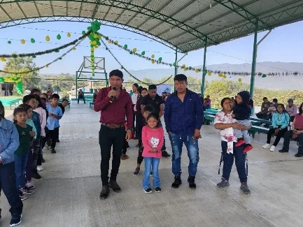 maestro de ceremonias con micrófono, niños y familias reunidas bajo palapa decorada