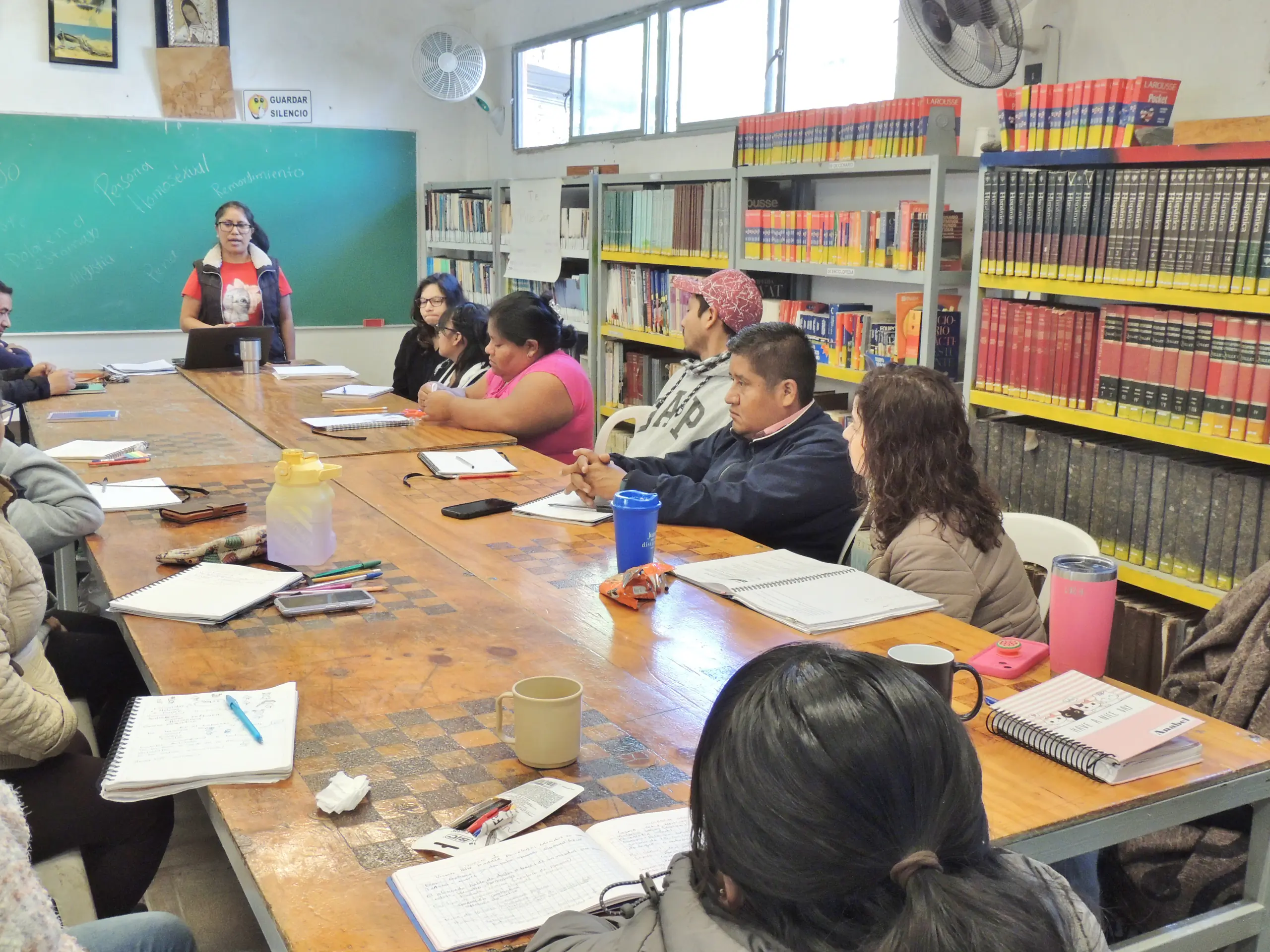 Docente y alumnos reunidos en la mesa grande de la biblioteca, con libros al fondo