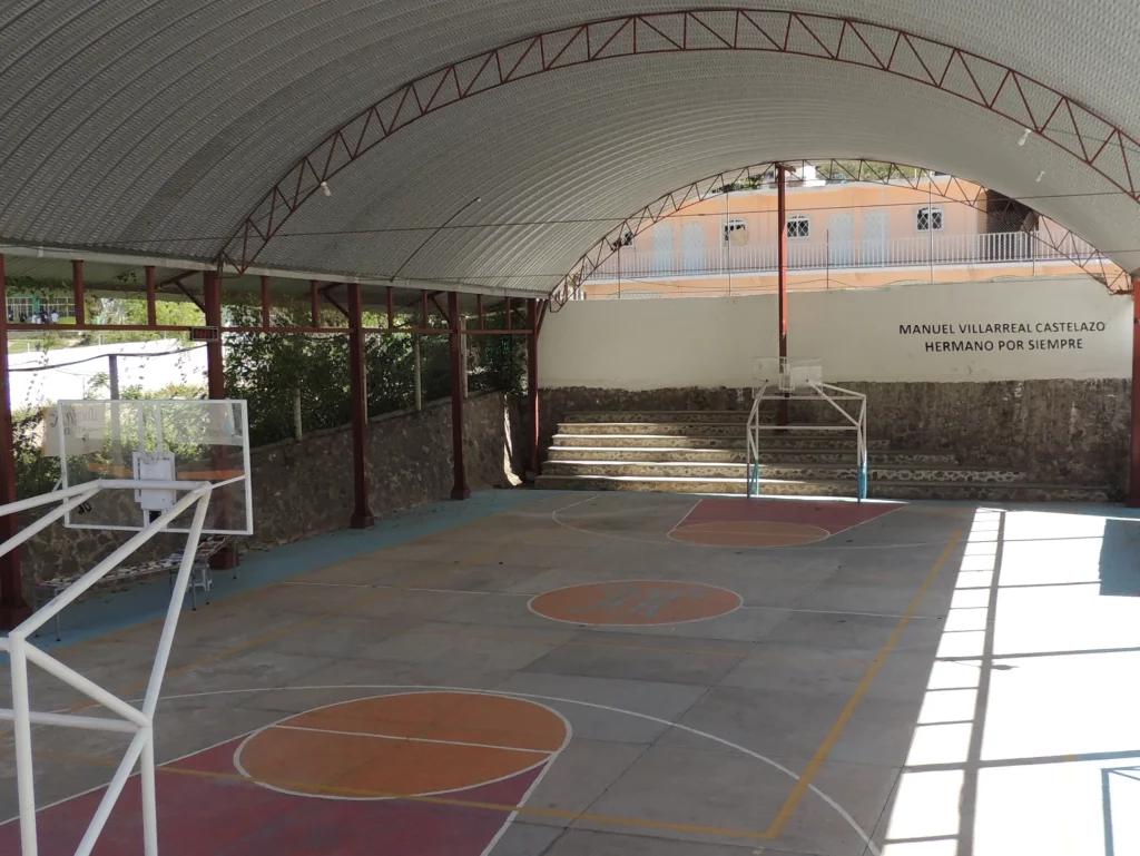 Cancha techada con arcos metálicos, tableros de básquetbol y gradas de piedra al fondo