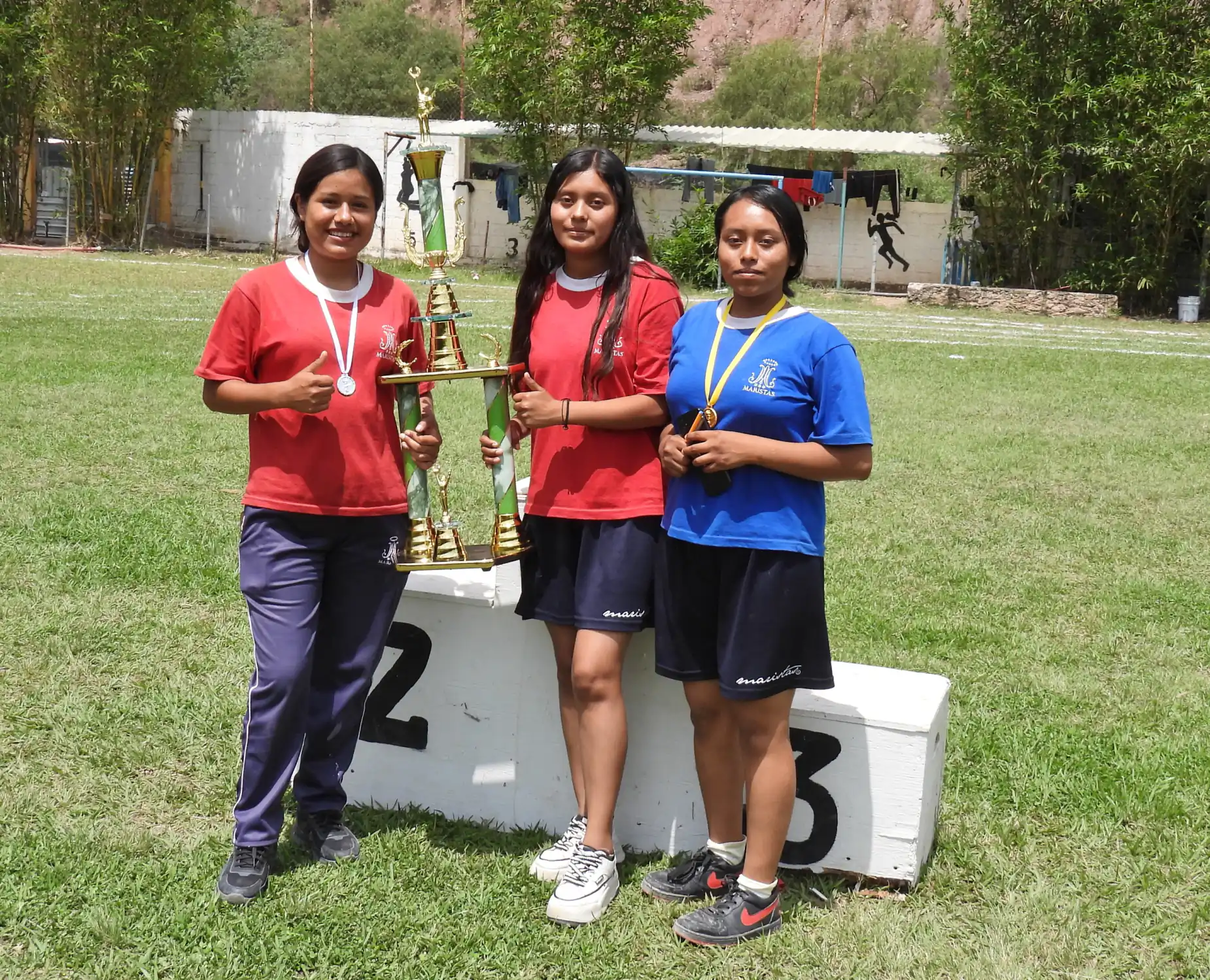 Alumnos con playera roja y medallas posando junto a un trofeo sobre el podio