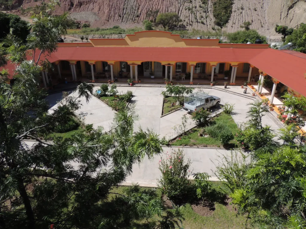 Patio con jardines y senderos y, al fondo, un edificio de arcos con techo de teja roja y la barranca