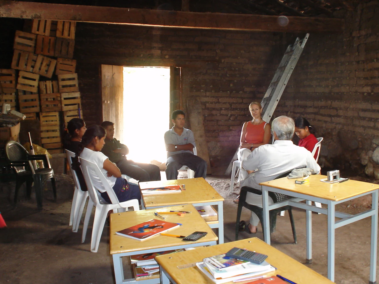 Alumnos y adultos sentados con mesas y libros en un salón de paredes de ladrillo con cajas de madera apiladas