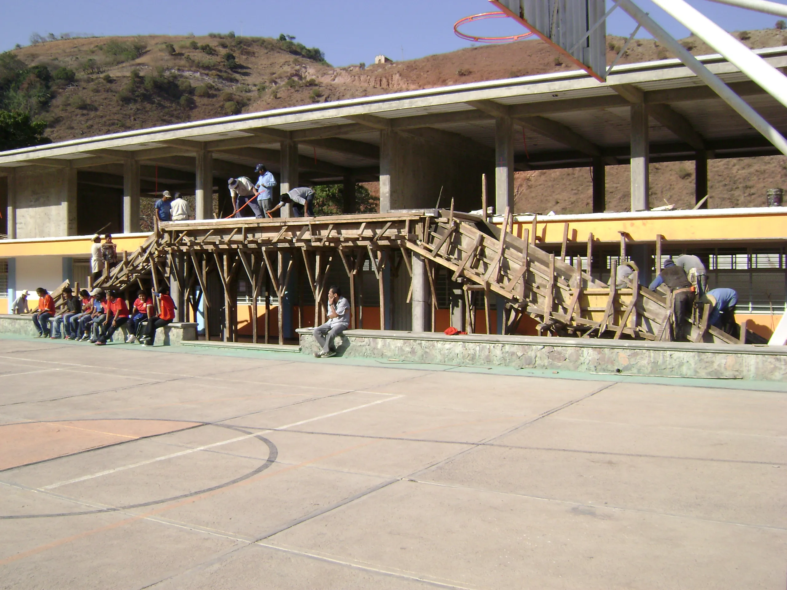 El edificio de la escuela en obra, con la estructura del segundo nivel y andamios de madera junto a la cancha