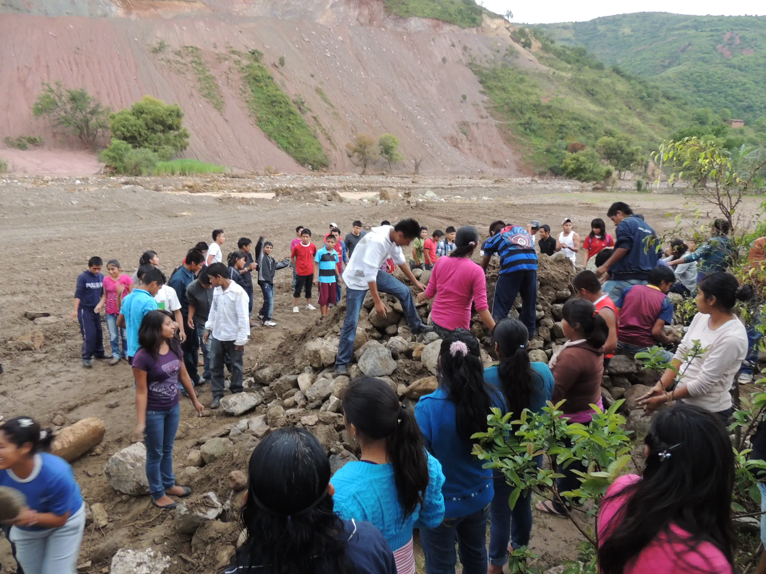 Decenas de estudiantes y adultos retirando piedras y lodo de un terreno arrasado por el río, con un cerro de tierra roja al fondo